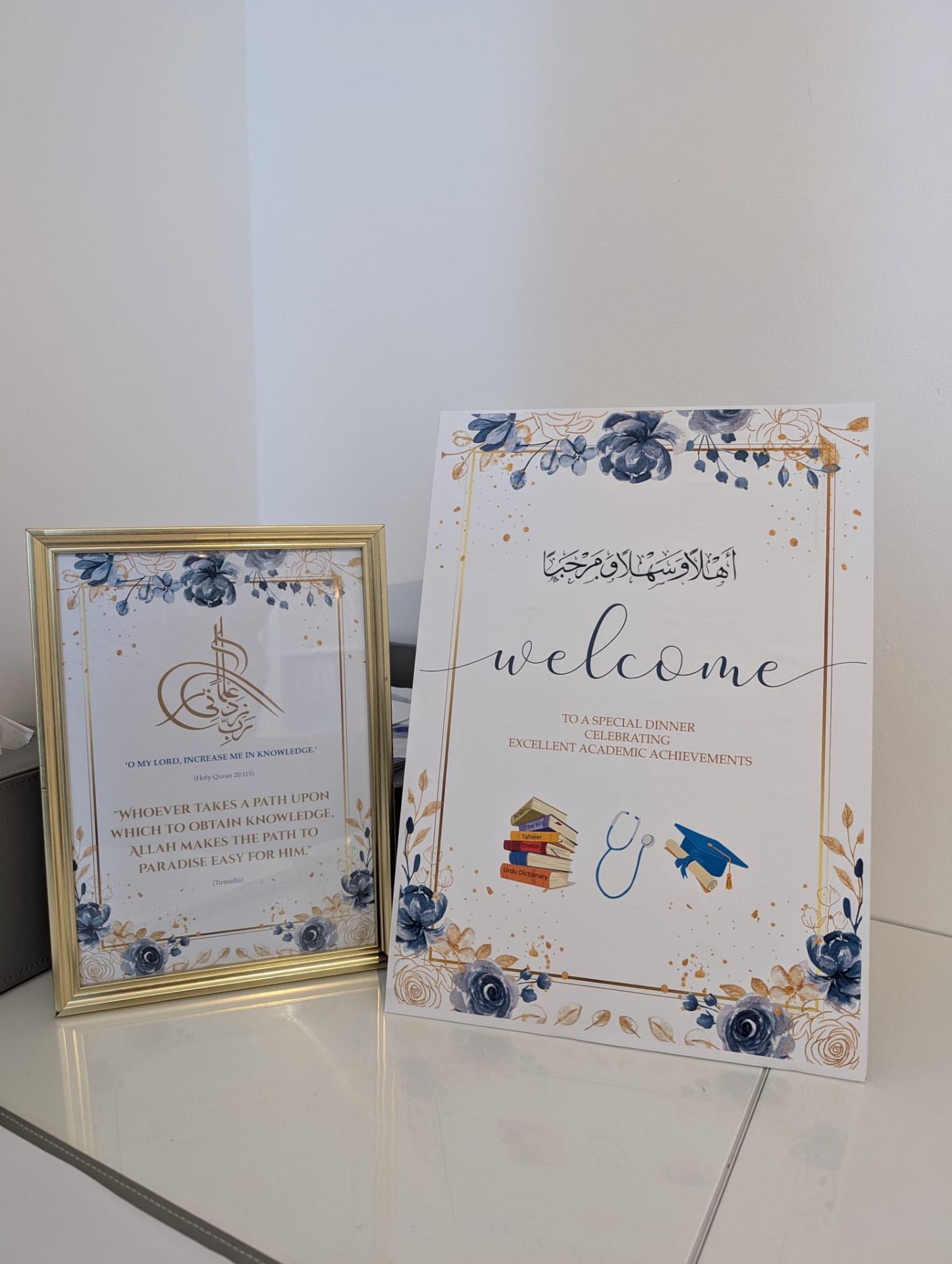 A blue and gold floral welcome sign for an academic achievements dinner, beside a gold-framed print with the dua for knowledge